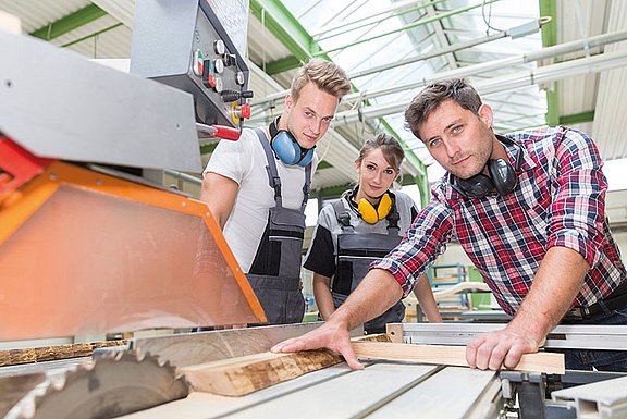 Foto: Eine Tischler zeigt zwei Tischlerlehrlingen, wie man an einer Formatkreissäge Holz sägt. Alle sind in Arbeitskleidung und haben einen Gehörschutz.