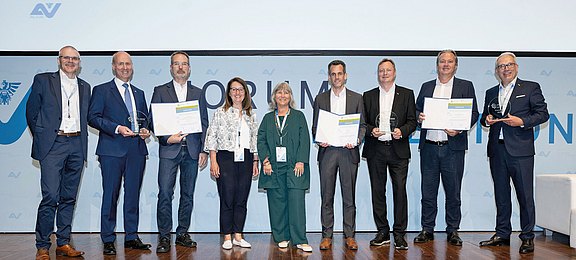 Foto: Mehrere Preisträgerinnen und Preisträger stehen gemeinsam auf einer Bühne und präsentieren Urkunden sowie Auszeichnungen. Die feierliche Gruppenaufnahme dokumentiert eine Preisverleihung im Rahmen einer professionell organisierten Fachveranstaltung. 