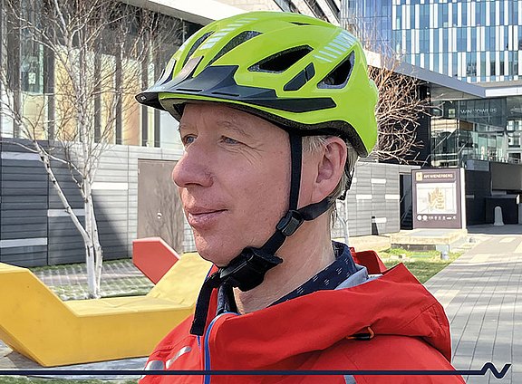 Foto: Eine Person trägt einen auffälligen neongrünen Fahrradhelm und eine rote Jacke im städtischen Umfeld. Die Nahaufnahme veranschaulicht korrektes Sicherheitsverhalten beim Radfahren und betont Sichtbarkeit im Straßenverkehr.