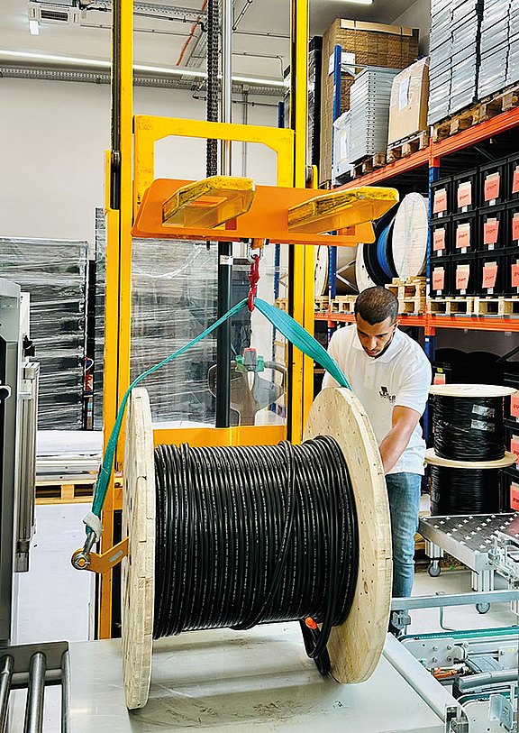 In einem Lagerhaus hantiert ein Arbeiter mit einer großen Kabeltrommel, die von einem gelben Gabelstapler gehalten wird. Er befestigt ein grünes Gurtband, um die Trommel zu sichern. Im Hintergrund sind Regale mit Lagerbeständen zu sehen, was auf eine geschäftige industrielle Umgebung hindeutet.