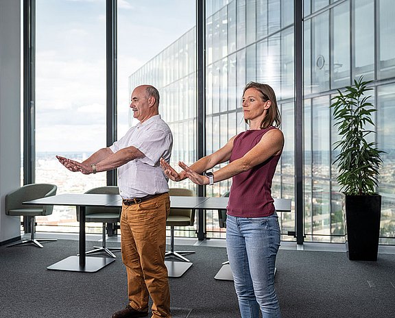 Foto: Zwei Personen demonstrieren eine Atemübung mit nach vorne ausgestreckten Armen. Die kontrollierte Bewegung fördert bewusste Atmung, Mobilität und Entspannung in einem modernen Büro mit weiter Aussicht.