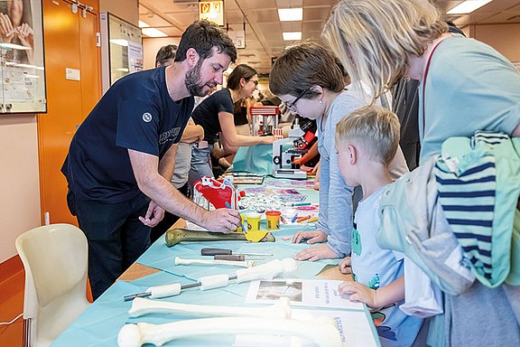 Foto: Ein Mann erklärt Kindern an einem Tisch mit Anatomie-Modellen etwas und demonstriert mit einem Werkzeug an einem großen Knochen.