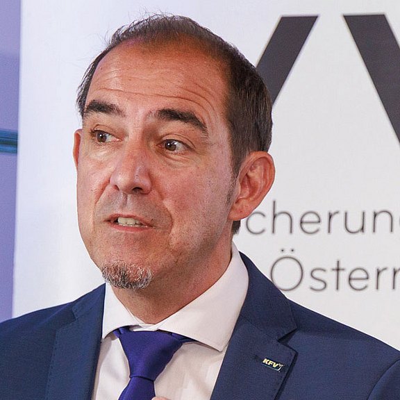 Foto: Ein Redner im blauen Anzug präsentiert bei einer Pressekonferenz des Versicherungsverbands Österreich. Mit ausdrucksstarker Gestik spricht er am Stehtisch, während Banner, Namensschilder und Wassergläser den Rahmen bilden.