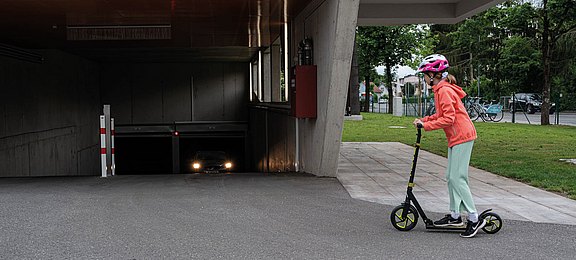Auf Garagenausfahrten achten! Kind mit Helm auf einem Roller vor einer Garagenauffahrt, in der gearde ein Auto aus der dunklen Ausfahrt fährt.