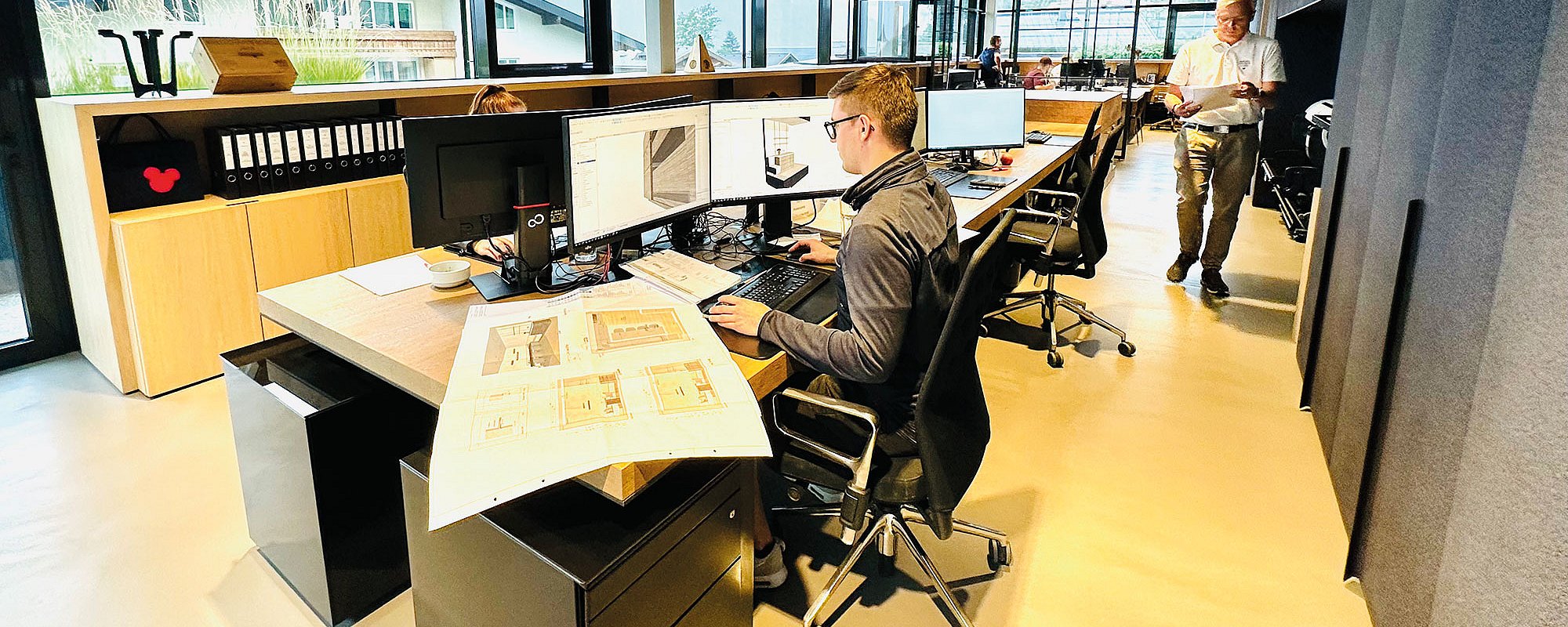 Großraumbüro mit Fensterfront und offenen Schreibtischplätzen mit hellem Boden und schwarzen Schreibtischen mit Holzplatte. Mitarbeiter arbeiten an den Computern mit Plänen auf den Schreibtischen.