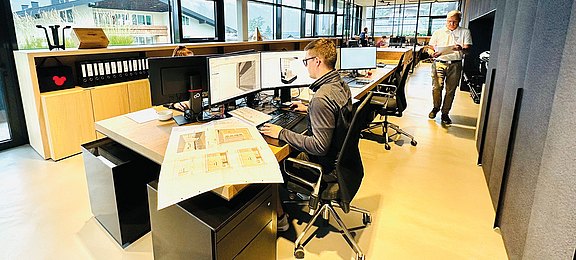 Großraumbüro mit Fensterfront und offenen Schreibtischplätzen mit hellem Boden und schwarzen Schreibtischen mit Holzplatte. Mitarbeiter arbeiten an den Computern mit Plänen auf den Schreibtischen. 