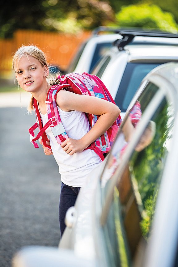 Foto: Ein blondes Mädchen mit einem bunten Schulranzen steht zwischen geparkten Autos und schaut, ob ein Auto kommt.