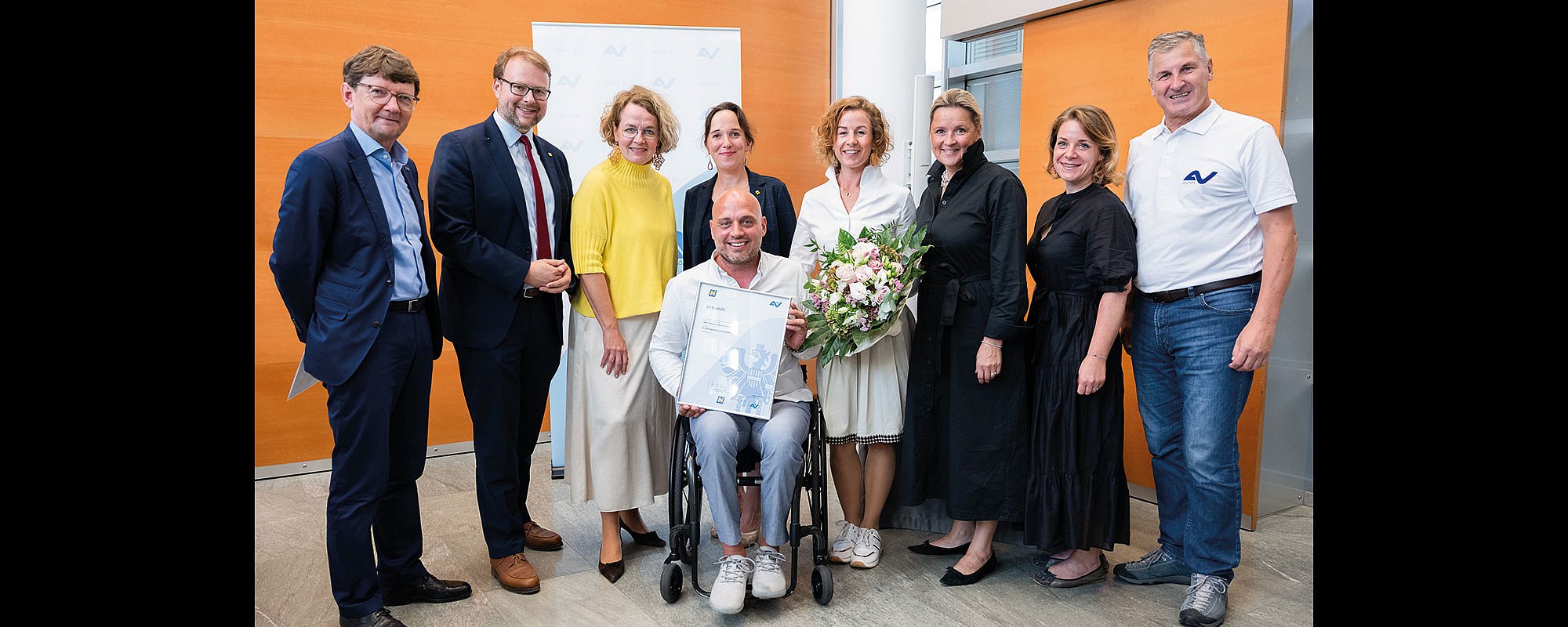 Gruppenbild mit fünf Frauen und vier Männern. Ein Mann im Rollstuhl hält eine gerahmte Auszeichnung ins Bild. Eine Dame daneben hält einen Blumenstrauß.