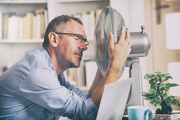 Foto: Ein Mann mit Brille sitzt am Schreibtisch und lehnt sein Gesicht mit geschlossenen Augen nah an einen Tischventilator, um sich abzukühlen.