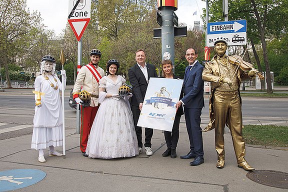 Mehrere verkleidete Personen posieren gemeinsam an einer Straßenkreuzung neben offiziellen Vertretern. Einige tragen historische Kostüme oder goldene Ganzkörperbemalung. Im Mittelpunkt steht ein Plakat, während Verkehrsschilder die urbane Umgebung markieren deutlich.