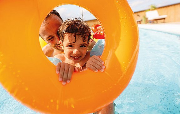 Foto: Ein lachender Junge mit nassen Haaren und Schwimmflügeln schaut mit seinem Vater durch einen orangen Schwimmring.