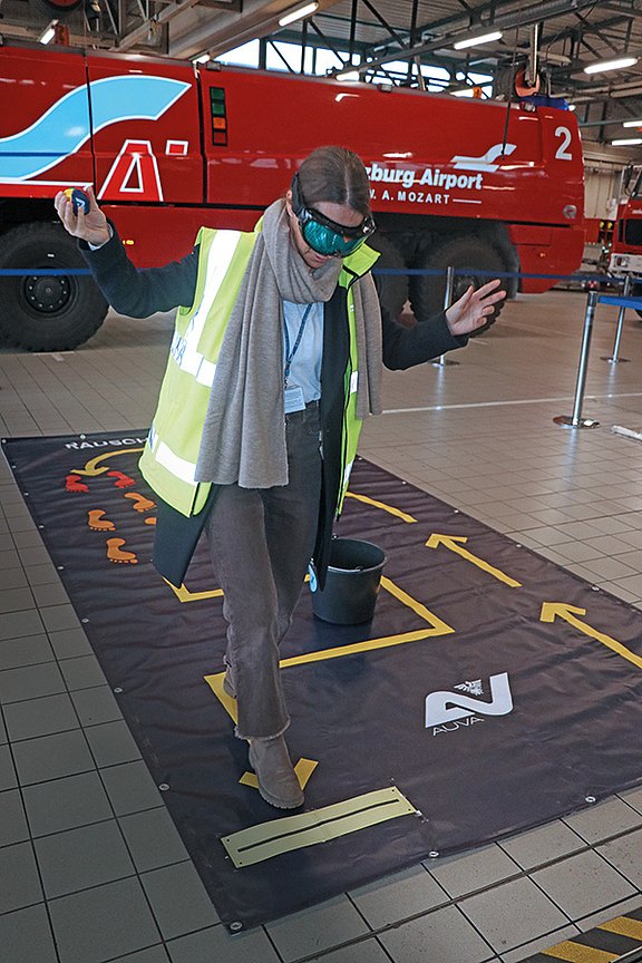 Das Bild zeigt eine Person mit reflektierender Sicherheitsweste und Rauschbrille, die auf einer Übungsmatte balanciert. Im Hintergrund steht ein rotes Flughafen-Feuerwehrfahrzeug mit der Aufschrift „Salzburg Airport W. A. Mozart“.