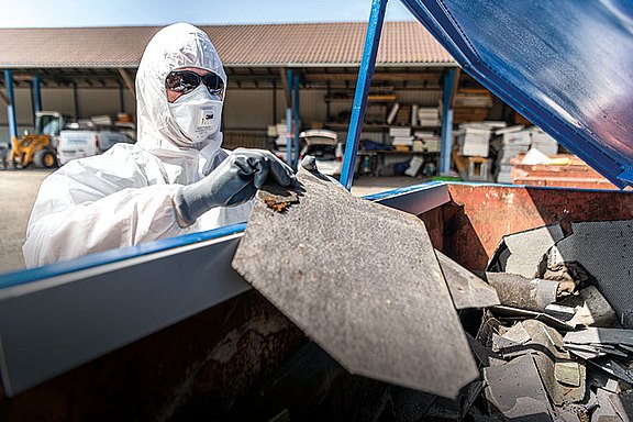 Foto: Eine Person trägt vollständige Schutzkleidung und entsorgt asbesthaltiges Material in einem Container. Die Aufnahme verdeutlicht fachgerechten Umgang mit Gefahrstoffen sowie konsequente Einhaltung von Sicherheits- und Arbeitsschutzmaßnahmen.