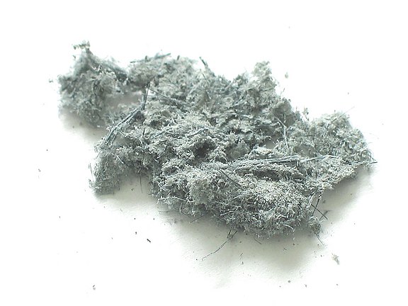 Auf hellem Hintergrund liegt eine graue, faserige Materialprobe mit feinen, verfilzten Strukturen. Die Nahaufnahme zeigt deutlich die unregelmäßige Oberfläche und die typischen Fasern eines mineralischen oder industriellen Stoffes.