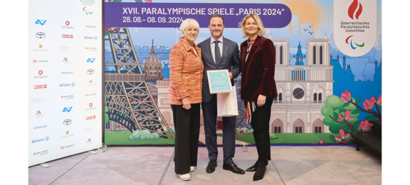 Das Bild zeigt drei Personen vor einem bunten Hintergrund mit dem Schriftzug „XVII. Paralympische Spiele, Paris 2024”. Der Mann in der Mitte, flankiert von zwei Frauen, hält eine Urkunde und eine Geschenktüte. Alle drei lächeln und stehen in offizieller pose.