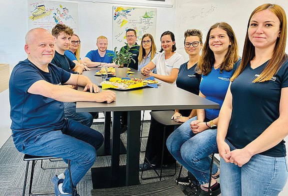 Eine Gruppe von etwa zehn Personen sitzt um einen Tisch in einem hellen Büro. Auf dem Tisch steht ein Teller mit Snacks und Getränken. Einige der Personen tragen identische T-Shirts mit einem Firmenlogo, was auf ein informelles Teamtreffen oder eine Feier hindeutet. Im Hintergrund sind bunte Zeichnungen an der Wand zu sehen.