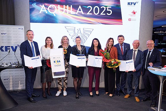 Das Foto zeigt eine feierliche Preisverleihung bei der Veranstaltung „AQUILA 2025“. Mehrere Personen stehen auf einer Bühne, halten Urkunden, Trophäen und Blumen. Im Hintergrund sind Logos des KFV und weiterer Institutionen zu sehen.