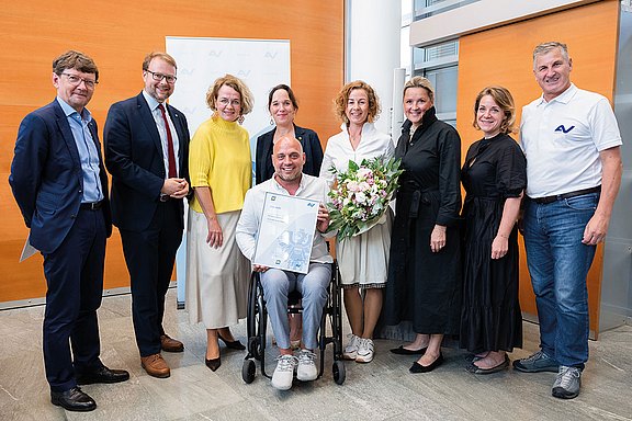 Gruppenbild mit fünf Frauen und vier Männern. Ein Mann im Rollstuhl hält eine gerahmte Auszeichnung ins Bild. Eine Dame daneben hält einen Blumenstrauß.