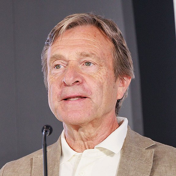 Foto: Ein Experte spricht bei einer Pressekonferenz an einem Stehtisch mit Mikrofon. Gestik, Namensschild und Wasserkaraffe unterstreichen den professionellen Veranstaltungscharakter vor Publikum.