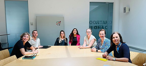 Foto: Sieben Personen sitzen gemeinsam an einem Besprechungstisch in einem hellen Konferenzraum. Die freundliche Gruppenaufnahme vermittelt Zusammenarbeit, Austausch und Teamgeist in einer professionellen Arbeitsatmosphäre mit moderner Büroeinrichtung. 