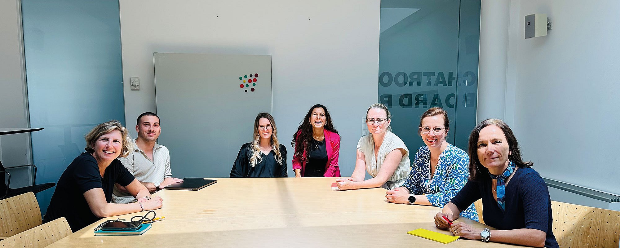 Foto: Sieben Personen sitzen gemeinsam an einem Besprechungstisch in einem hellen Konferenzraum. Die freundliche Gruppenaufnahme vermittelt Zusammenarbeit, Austausch und Teamgeist in einer professionellen Arbeitsatmosphäre mit moderner Büroeinrichtung.